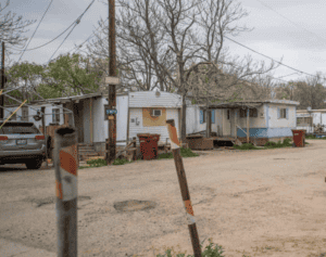 Trailer homes on a dirt road with trees and a parked car, under a cloudy sky.